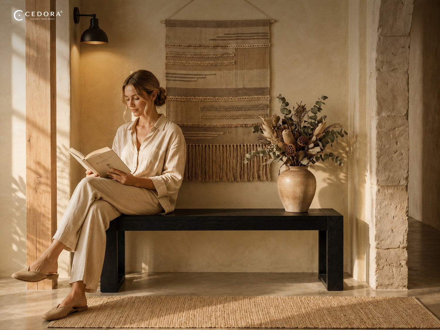 Woman reading on a black timber bench beside a stoneware vase of dried foliage and a woven wall hanging in a sun lit hallway