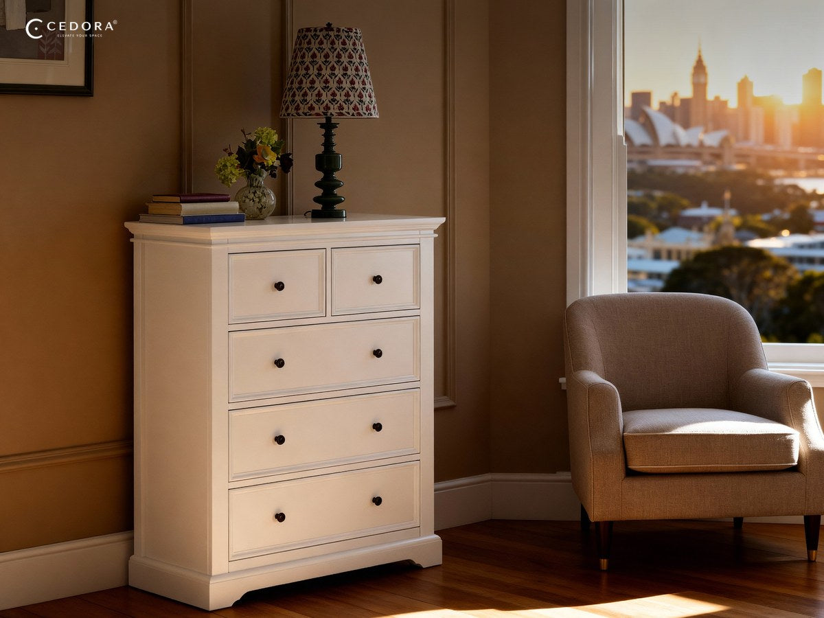 Tall white five drawer tallboy against a dark panelled wall beside an armchair and a window at sunset