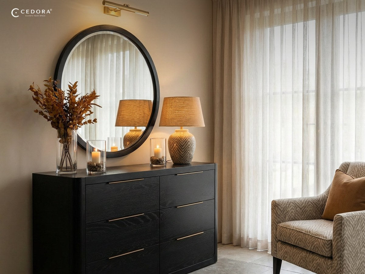 Round black framed mirror above a black six drawer chest with lamps and candles in warm afternoon light