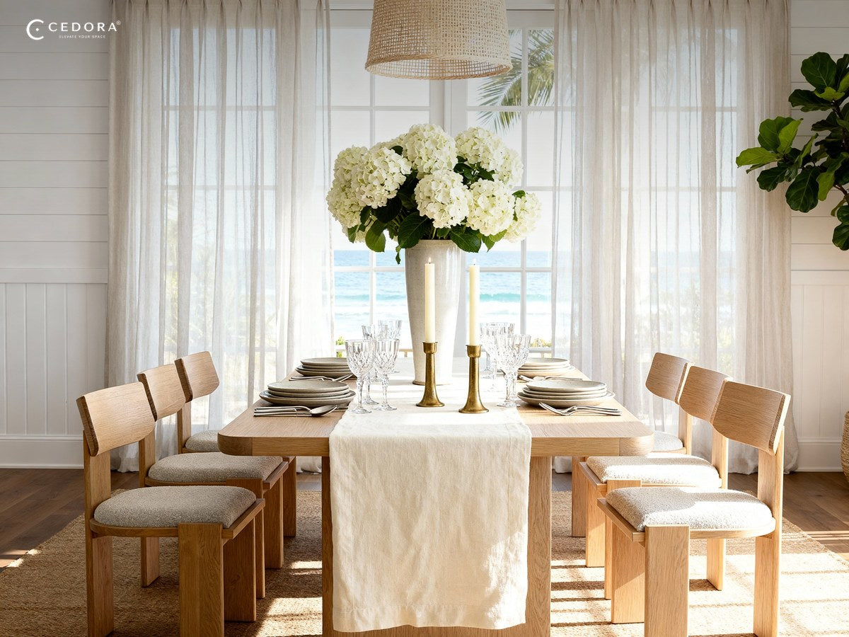 Natural timber dining table set with candles and hydrangeas in front of sheer curtains and an ocean view