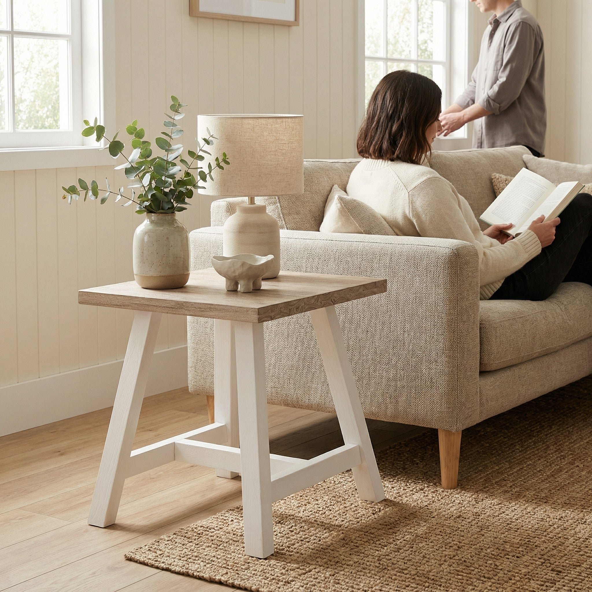 Woman reading a book on a beige sofa in a cozy living room with a wooden side table and decorative items.

