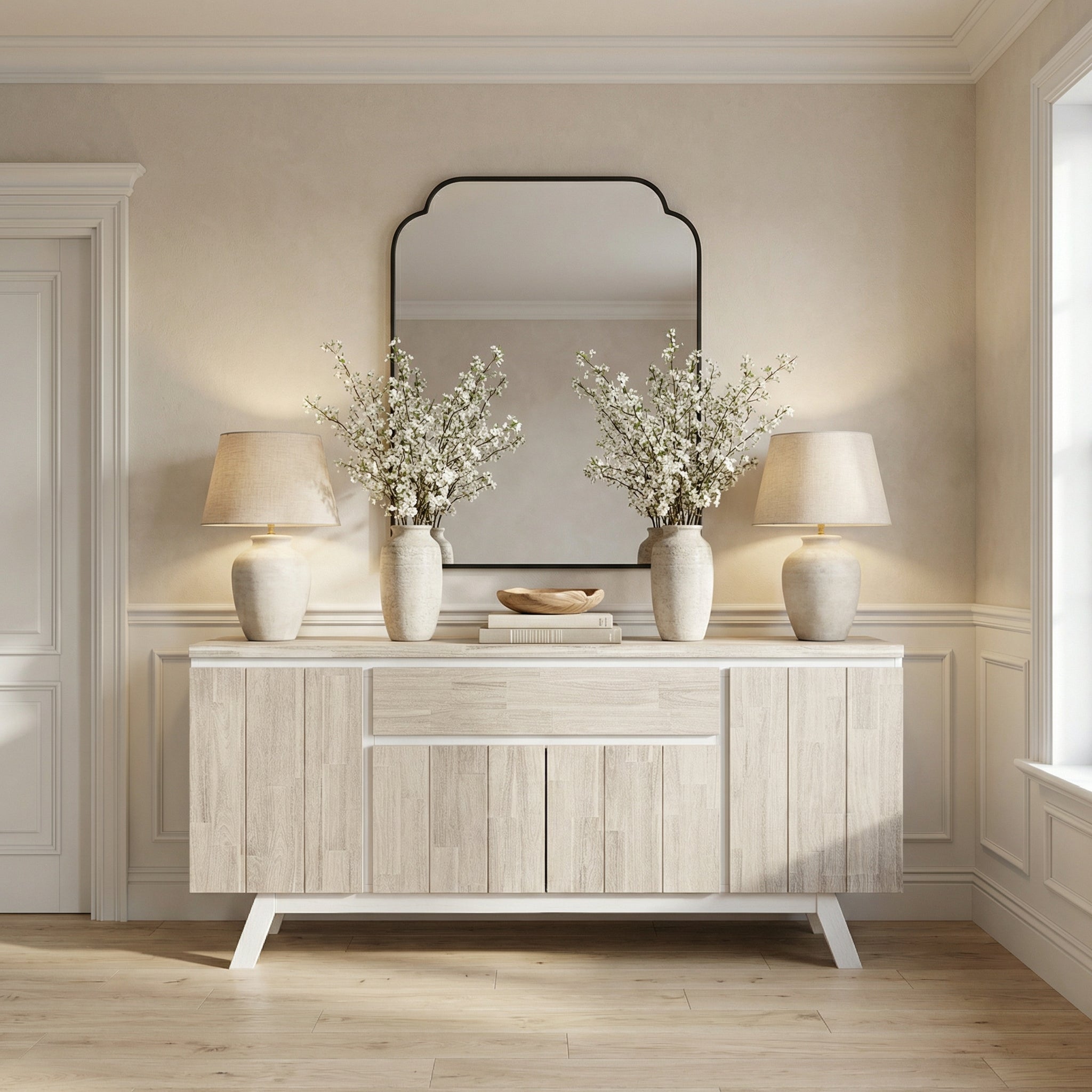 Light wood sideboard with two lamps, two vases with white flowers, and a mirror in a room.

