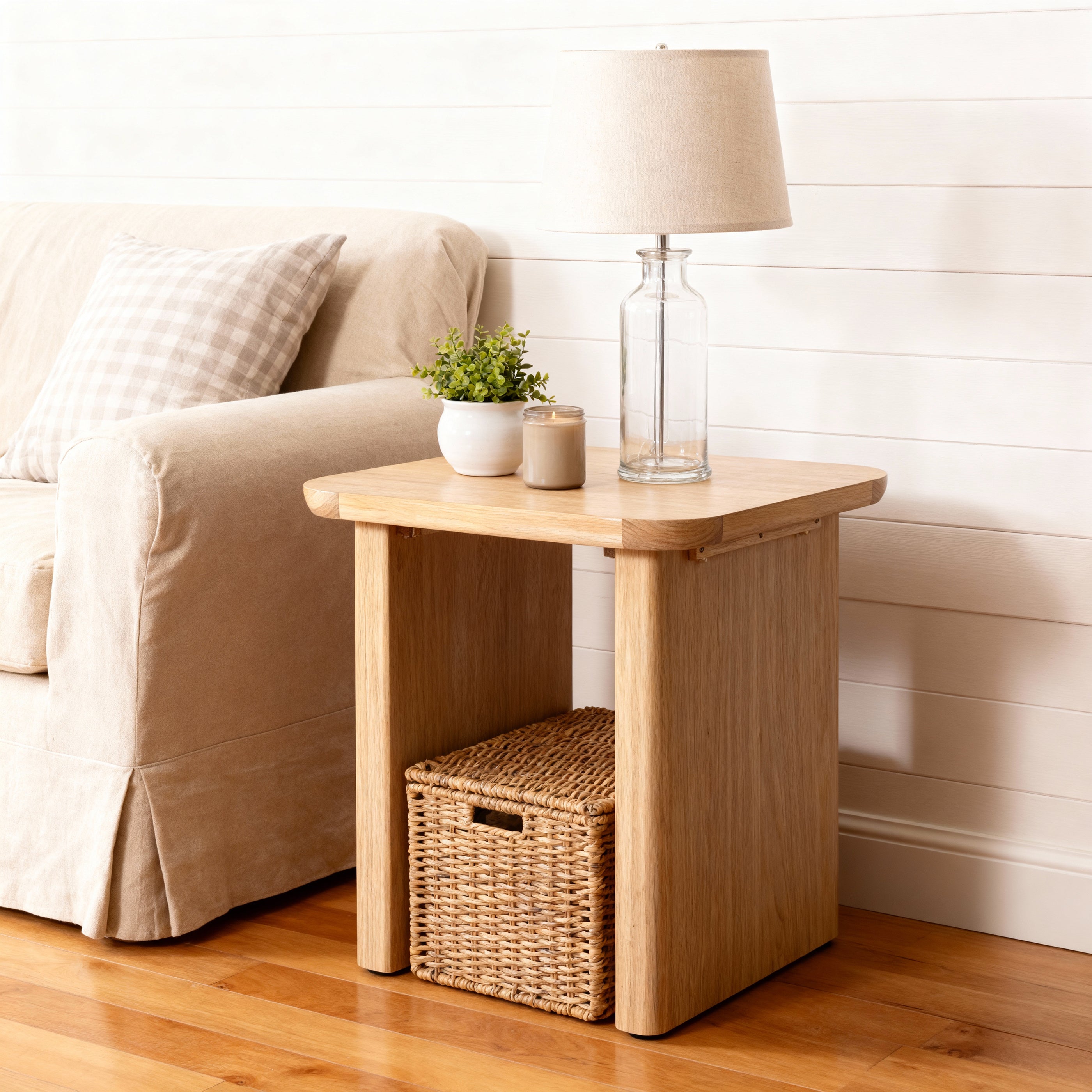 Wooden side table with a lamp, plant, and basket next to a beige sofa.

