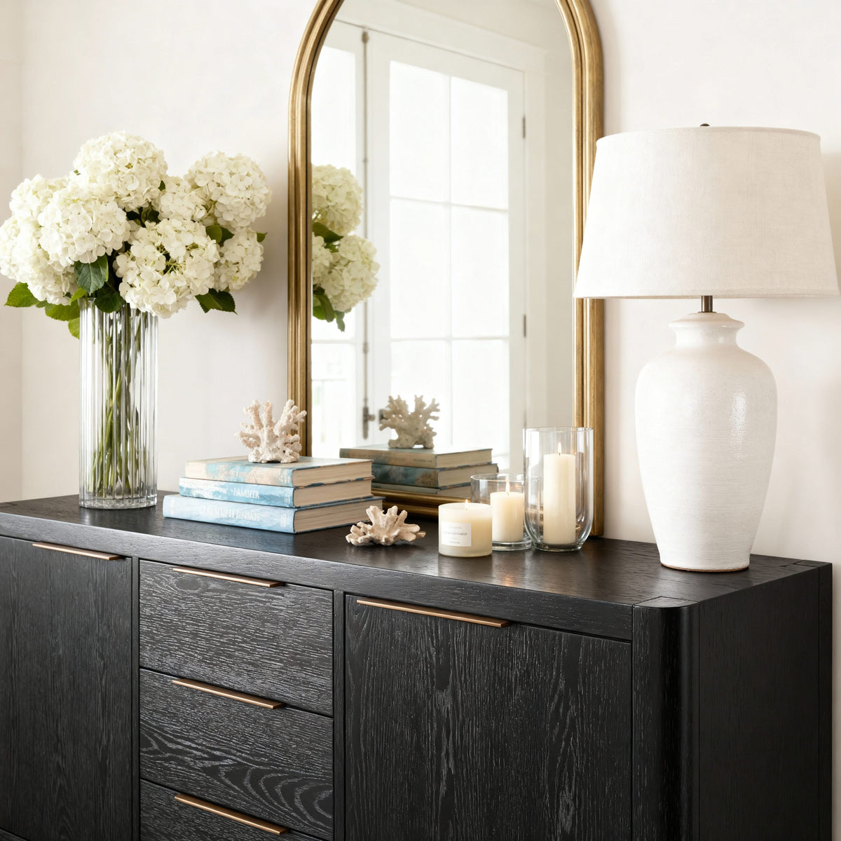 Decorative setup with a vase of white flowers, books, candles, and a lamp on a dark wooden dresser.

