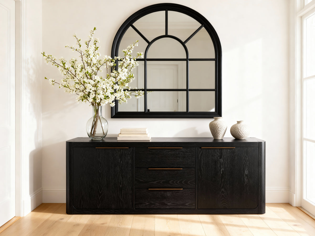 Black dresser with mirror, flowers, and decorative items in a bright room.

