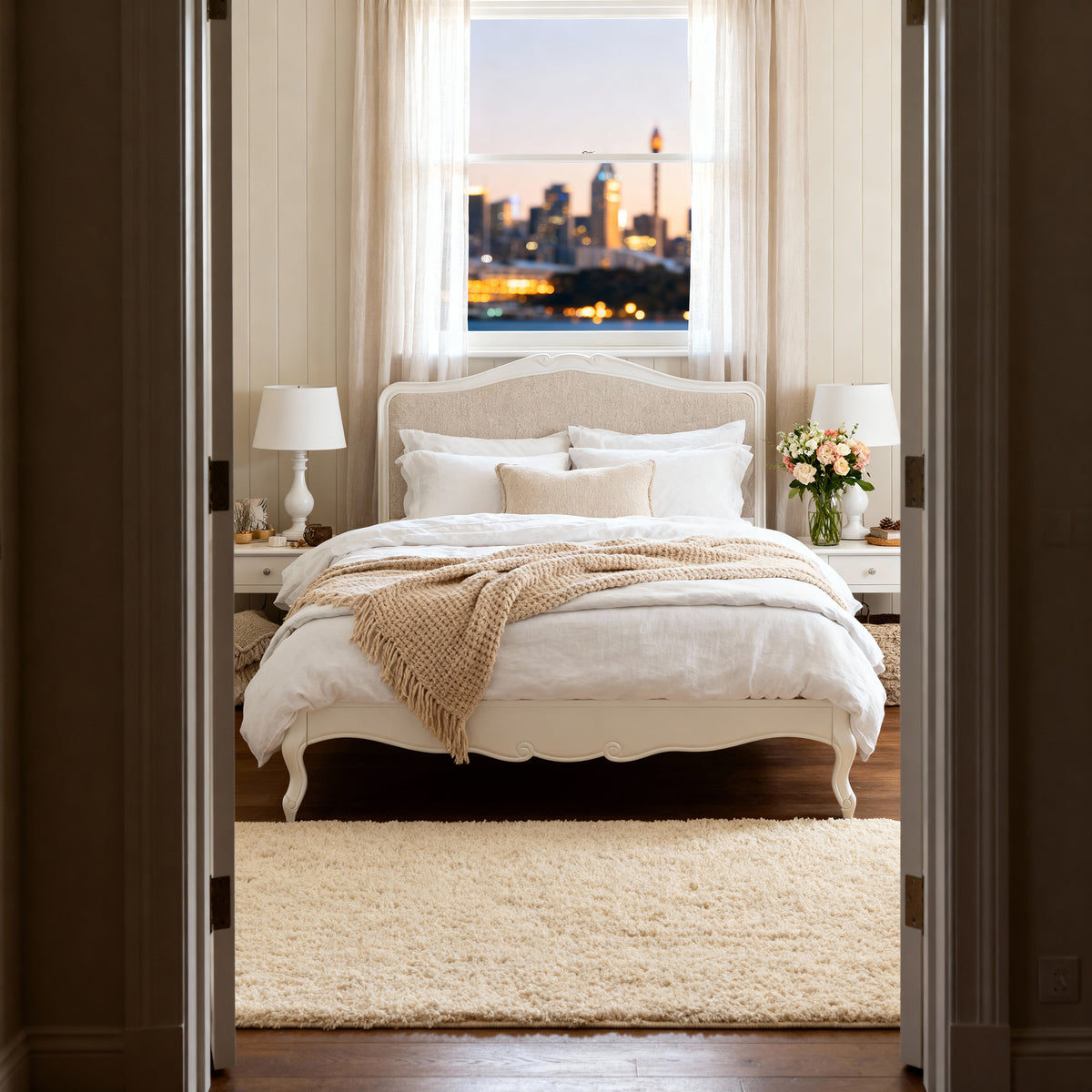 Newcastle Acacia Queen Bed Frame seen through open double doors, dressed in cream linen by a window at dusk
