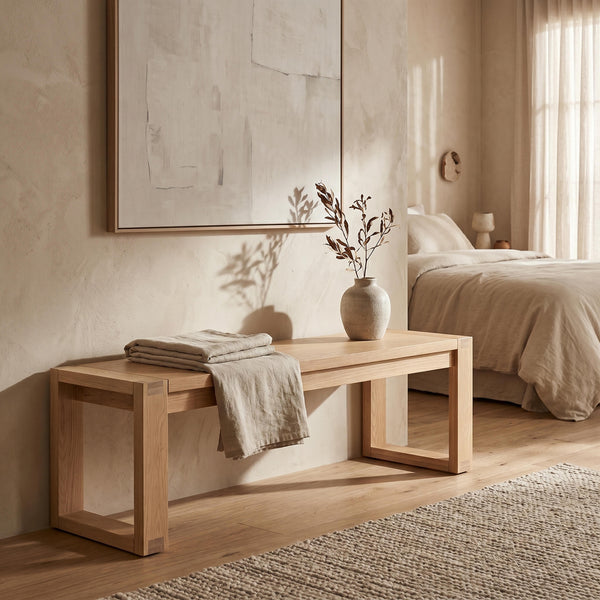 Wooden bench with folded towels in a bedroom setting with a bed and abstract art on the wall.

